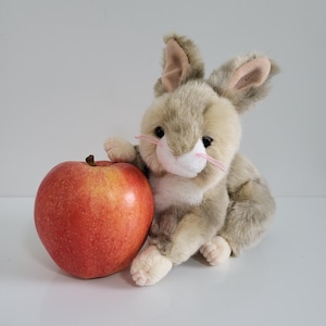 May include: A plush gray and white bunny rabbit toy sitting next to a red apple. The bunny has long ears and a white nose.