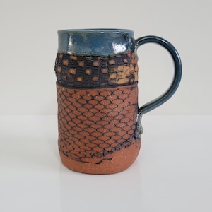 May include: A handmade ceramic mug with a blue glaze and a textured brown body. The mug has a unique design with a woven pattern and a textured surface.