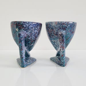 May include: Two speckled ceramic mugs with a unique design. The mugs are blue with red and teal accents, featuring a handle and a triangular base. The mugs have a speckled pattern and a glossy finish.