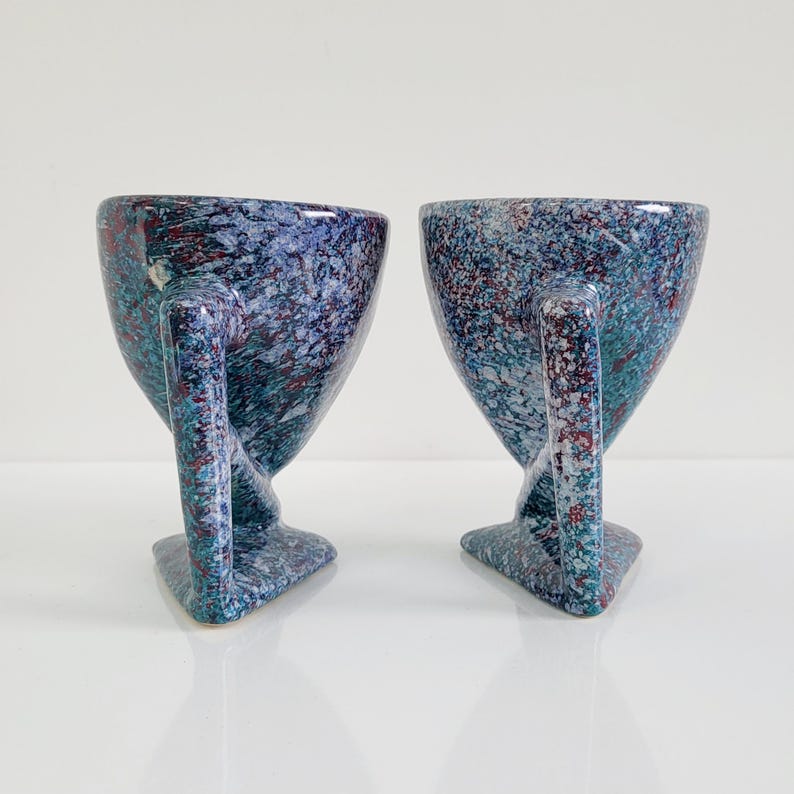 May include: Two speckled ceramic mugs with a unique design. The mugs are blue with red and teal accents, featuring a handle and a triangular base. The mugs have a speckled pattern and a glossy finish.