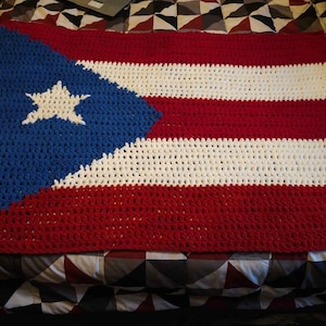 May include: A crocheted blanket featuring the flag of Puerto Rico. The design includes a blue triangle with a white star, and alternating red and white horizontal stripes. The blanket is handmade and appears to be soft and cozy.