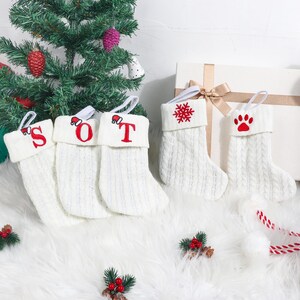 Peut inclure: Chaussettes de Noël en tricot blanc avec des détails brodés en rouge. Les bas de Noël présentent les lettres "SOT", un flocon de neige et une empreinte de patte. Chaque bas a une boucle blanche pour l'accrocher. Un sapin de Noël est en arrière-plan.