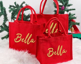 Könnte beinhalten: Drei rote Tragetaschen mit goldglitzernden Buchstaben, die "Bella" buchstabieren. Die Taschen sind unterschiedlich groß und haben passende rote Griffe. Die Taschen stehen auf einer weißen, flauschigen Oberfläche, mit einem Weihnachtsbaum im Hintergrund.
