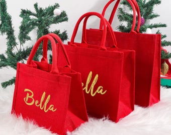 Könnte beinhalten: Drei rote Jute-Tragetaschen in verschiedenen Größen mit goldenem Schriftzug "Bella". Die Taschen haben passende rote Griffe und sind auf einer weißen Oberfläche mit einem Weihnachtsbaum im Hintergrund platziert.
