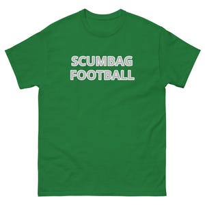 Może przedstawiać: Zielona koszulka z napisem "SCUMBAG FOOTBALL" w białych literach z szarym obrysem. Koszulka jest jednokolorowa i ma krótkie rękawy.