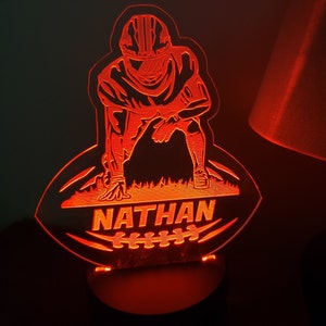 May include: Red illuminated night light shaped like a football with a football player in a three-point stance. The name "NATHAN" is written on the football.