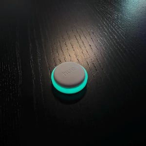May include: A small, circular, grey device with the word "hue" printed on its top surface. It features a glowing, turquoise-coloured ring around the edge. The device is on a dark wooden surface.