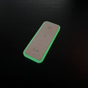 Op de afbeelding: Een rechthoekige afstandsbediening met een lichtgroene rand en een grijze voorkant. De afstandsbediening heeft drie knoppen met lichtsymbolen en het woord "hue". De bovenste knop heeft een aan/uit-symbool.