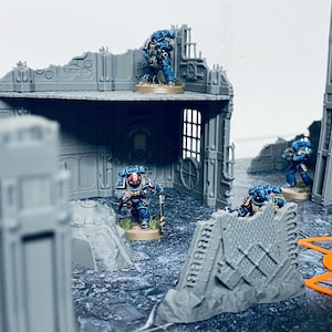 Könnte beinhalten: Eine Miniatur-Krieger-Szene mit blauen Space Marine-Figuren in einer zerstörten Stadtlandschaft. Die Figuren sind in verschiedenen Kampfstellungen mit gezogenen Waffen positioniert. Die Szene spielt auf einer grauen und blauen, strukturierten Oberfläche.