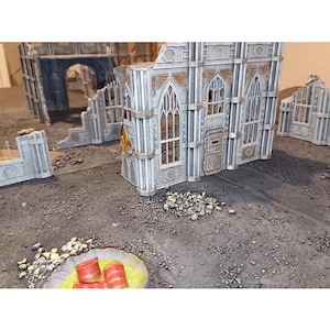 Puede incluir: Un modelo de un edificio en ruinas con paredes grises y ventanas arqueadas. El edificio está hecho de plástico y es parte de un juego de guerra de mesa. El edificio está sentado sobre una superficie gris con pequeñas piedras y una pequeña pila de barriles rojos.