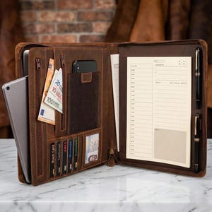 Könnte beinhalten: Eine braune Ledermappe, geöffnet, um verschiedene Fächer zu zeigen. Sie enthält ein Tablet, Bargeld, Kreditkarten, ein Smartphone und einen Notizblock mit zwei Stiften. Die Mappe ist mit einem Reißverschluss versehen und liegt auf einer Marmoroberfläche.