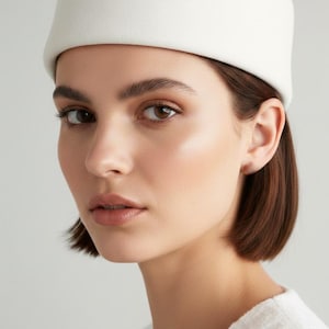 Può includere: Un cappello bianco in stile pillbox con una corona arrotondata. Il cappello è realizzato in un materiale morbido e strutturato. La modella ha i capelli corti e castano scuro e indossa un top bianco. Lo sfondo è grigio chiaro.