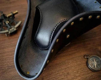 Pode incluir: Um chapéu tricórnio de couro preto com uma copa lisa e arredondada e uma aba larga virada para cima. As bordas da aba são reforçadas com costuras visíveis e acentuadas com pequenos rebites metálicos de cor latão espaçados uniformemente.