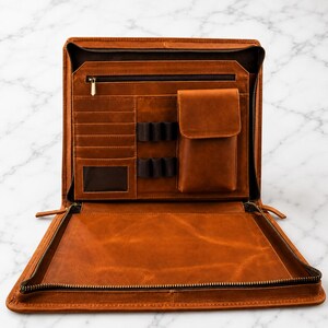 Könnte beinhalten: Geöffnetes, braunes Leder-Portfolio mit mehreren Fächern, darunter Kartenfächer, Stifthalter und eine kleine Tasche. Das Portfolio hat einen Reißverschluss und eine transparente Tasche. Der Innenraum ist mit dunklerem braunen Material ausgekleidet.