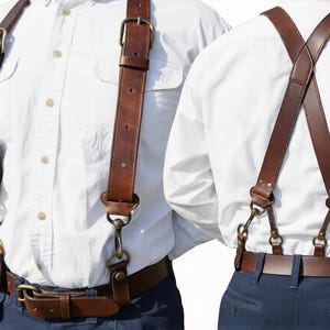 May include: Brown leather suspenders with brass-coloured hardware. The suspenders are attached to a brown leather belt. The suspenders are worn over a white button-down shirt and blue trousers. The suspenders have adjustable straps.