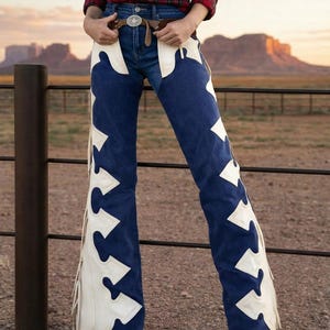 May include: Blue denim chaps with white decorative accents and fringe. The chaps are worn over blue jeans and paired with a brown belt and a red plaid shirt. The background features a desert landscape.