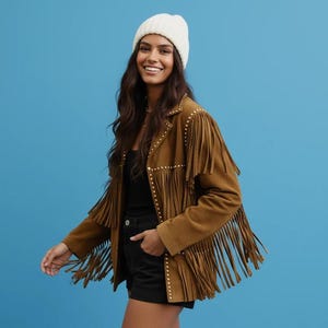 Könnte beinhalten: Braune Wildlederjacke mit Fransen und goldenen Nieten. Die Jacke wird mit einer weißen Mütze, einem schwarzen Oberteil und schwarzen Shorts kombiniert. Der Hintergrund ist einfarbig blau.