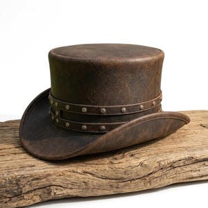 Peut inclure: Un haut-de-forme marron foncé avec une texture vieillie et usée. Le chapeau présente un bord large et incurvé ainsi qu'une bande à double épaisseur autour de la calotte, ornée de petits clous métalliques régulièrement espacés sur tout le pourtour.