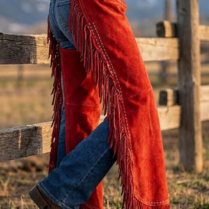 Puede incluir: Chaps de ante rojo con largos flecos, usados sobre vaqueros azules. Los chaps se aseguran con un cinturón en la cintura. Botas de vaquero marrones completan el look de inspiración western. La imagen está tomada al aire libre con una valla de madera al fondo.