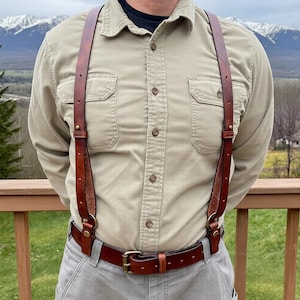 May include: Brown leather suspenders with brass fittings, worn over a tan button-up shirt and grey trousers. The suspenders are attached to a matching brown leather belt with a brass buckle.
