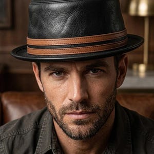 May include: A black leather fedora hat featuring a textured surface and a double-layered tan leather band wrapped around the base of the crown. The hat has a structured brim and a flat top, showcasing a classic design with visible stitching details.
