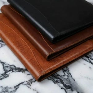 Könnte beinhalten: Drei Leder-Portfolios in Braun- und Schwarztönen sind auf einer Marmoroberfläche gestapelt. Das oberste Portfolio ist schwarz, das mittlere ist mittelbraun und das untere ist hellbraun. Die Portfolios haben ein glattes, professionelles Aussehen.