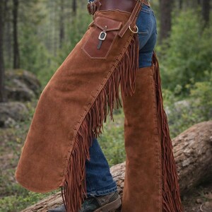 May include: Brown suede chaps with long fringe and a leather belt with a pocket. The chaps are worn over blue jeans and boots. The chaps are a western wear item.