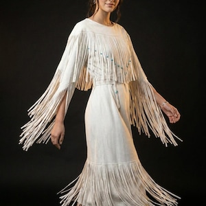Puede incluir: Vestido color crema con capa y falda con flecos. La capa presenta flecos largos y pequeñas cuentas turquesas. El vestido tiene cintura entallada y falda midi, también con flecos. El conjunto es de tejido suave y fluido.