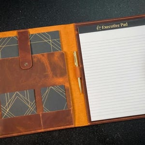 Könnte beinhalten: Geöffnetes Executive-Padfolio aus braunem und orangefarbenem Leder. Es hat einen linierten Notizblock, einen goldenen Stift und Taschen mit schwarzen Karten mit goldenen geometrischen Designs. Auf dem Notizblock steht "Executive Pad".