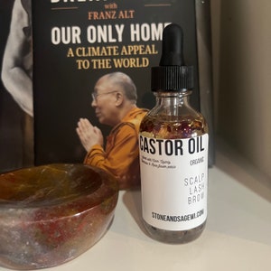 May include: A clear glass bottle with a black dropper top filled with a light yellow liquid and dried flowers. The bottle label reads "CASTOR OIL ORGANIC SCALP LASH BROW STONEANDSAGEWI.COM".