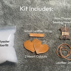 May include: A craft kit includes a bag of white polyester fiberfill, a small tan leather strap, two tan leather heart cutouts with pre-punched holes, a spool of wax thread with two sewing needles, and a length of tan leather string for assembly.