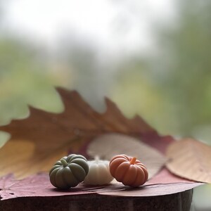 May include: Three small pumpkin-shaped candles, one green, one white, and one orange, are arranged on a brown leaf. The candles are on a wooden surface.