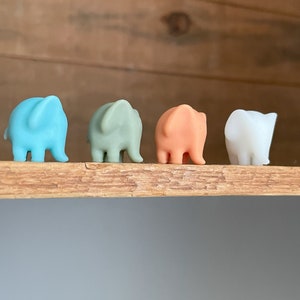Könnte beinhalten: Vier kleine Elefantenfiguren in Pastellfarben. Die Elefanten sind auf einem Holzregal in einer Reihe angeordnet. Die Farben sind hellblau, grün, pfirsichfarben und weiß. Der Hintergrund ist unscharf.