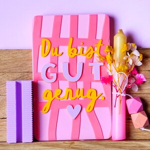 May include: A rectangular pink sign with a grid pattern and the yellow and lavender text "Du bist GUT genug." with a small heart. Beside it is a purple ribbed block, a pink candle with dried yellow and pink flowers, and two geometric beads.