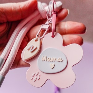 Puede incluir: Un llavero rosa en forma de flor con la palabra "Mama" en escritura blanca. El llavero tiene un pequeño corazón y una etiqueta en forma de hoja con la letra "N". Una correa rosa y un cierre transparente están adjuntos.