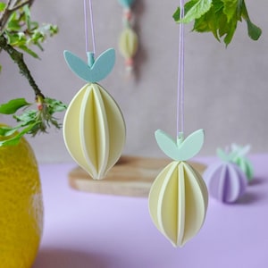 May include: Decorative paper ornaments in the shape of lemons, hanging from thin purple strings. The ornaments are yellow with light green leaf accents. Other ornaments in shades of blue, yellow, and purple are visible in the background. The background is a soft purple.