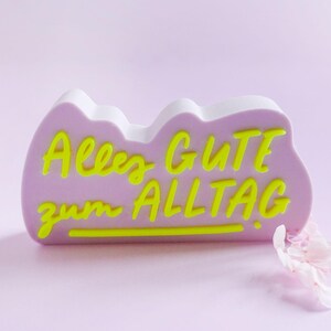 Può includere: Un oggetto decorativo color lavanda con il testo tedesco "Alles Gute zum Alltag" in giallo neon. L'oggetto ha una forma a nuvola con una parte superiore bianca. Alcuni petali rosa sono sullo sfondo su uno sfondo viola chiaro.