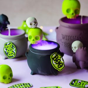 Puede incluir: Una colección de velas con forma de caldero en negro, gris y morado que emiten un brillo morado y humo. Las velas cuentan con calaveras de color verde lima, figuras de pájaros negros y etiquetas verde lima con el texto BOO y SPOOKY SEASON.