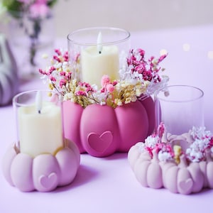 Puede incluir: Tres portavelas con forma de calabaza en tonos rosados, cada uno con un detalle en forma de corazón en la parte frontal. Cada soporte contiene un cilindro de cristal transparente con una vela blanca, rodeado de flores secas rosas y blancas.
