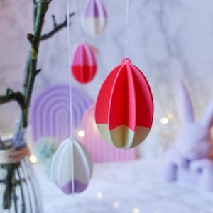 Peut inclure: Ornements d'œufs de Pâques suspendus en rouge, rose, blanc et violet. Les œufs sont suspendus à une branche avec un vase et un lapin en arrière-plan. Les œufs ont un design géométrique et sont peints avec une palette bicolore.