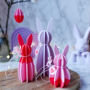 Könnte beinhalten: Eine Osterdekoration mit drei dekorativen Hasenfiguren in Rosa und Lila sowie hängenden Eierornamenten. Die Hasen sind mit Blumen und herzförmigen Anhängern verziert, auf einem Holzbrett platziert.