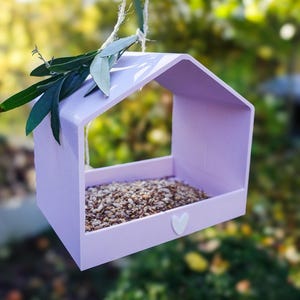 May include: A light purple bird feeder shaped like a house, filled with birdseed. A small white heart is on the front. Green leaves and a twine string are attached to the top. The feeder is hanging outdoors.