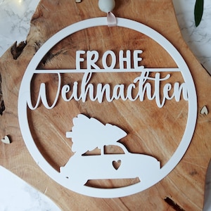 Könnte beinhalten: Weißes Weihnachtsornament aus Holz mit den Worten "FROHE Weihnachten" und einem Auto, das einen Weihnachtsbaum transportiert. Das runde Ornament hat ein Band zum Aufhängen. Der Hintergrund ist eine Holzoberfläche.