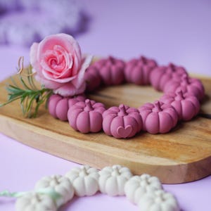 Op de afbeelding: Twee ronde kransen gemaakt van kleine, gevormde pompoenvormige kralen. De ene krans is donker mauve met een klein hartje op een pompoen en een roze roos bevestigd. De andere krans is gebroken wit en ligt op de voorgrond op een paars oppervlak.