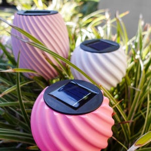 Puede incluir: Tres luces solares de jardín en rosa, lavanda y blanco, con un diseño en espiral. Cada luz tiene un panel solar negro en la parte superior y está colocada entre hierba verde y amarilla.