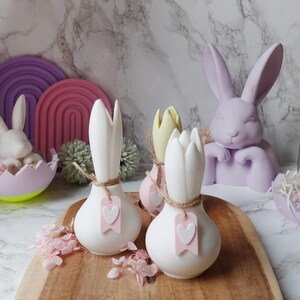 K&ouml;nnte beinhalten: Osterdekoration mit wei&szlig;en, zwiebelf&ouml;rmigen Vasen mit rosa Herzanh&auml;ngern, einem hellvioletten Hasen und anderen pastellfarbenen Osterdekorationen. Die Vasen sind mit Bindfaden gebunden. Die Artikel sind auf einem Holztablett angeordnet.