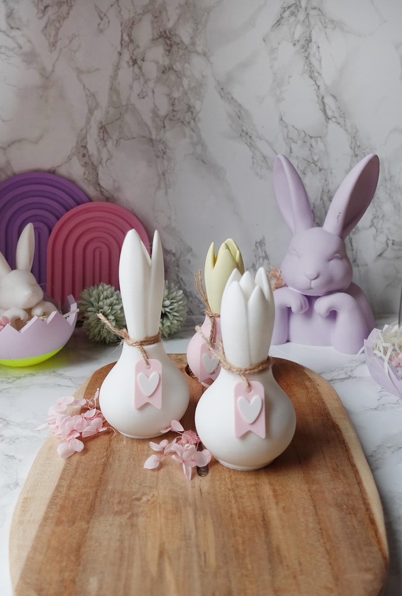 K&ouml;nnte beinhalten: Osterdekoration mit wei&szlig;en, zwiebelf&ouml;rmigen Vasen mit rosa Herzanh&auml;ngern, einem hellvioletten Hasen und anderen pastellfarbenen Osterdekorationen. Die Vasen sind mit Bindfaden gebunden. Die Artikel sind auf einem Holztablett angeordnet.