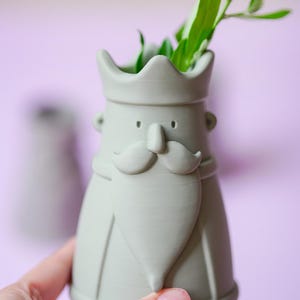 Peut inclure: Un vase en c&eacute;ramique vert sauge mat en forme de roi avec une couronne, une moustache et une barbe pointue. Le vase pr&eacute;sente une ouverture sup&eacute;rieure creuse contenant des feuilles vertes et une finition lisse avec des lignes d'impression 3D.