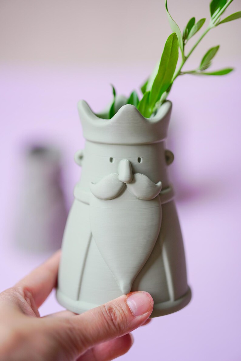 Peut inclure: Un vase en c&eacute;ramique vert sauge mat en forme de roi avec une couronne, une moustache et une barbe pointue. Le vase pr&eacute;sente une ouverture sup&eacute;rieure creuse contenant des feuilles vertes et une finition lisse avec des lignes d'impression 3D.