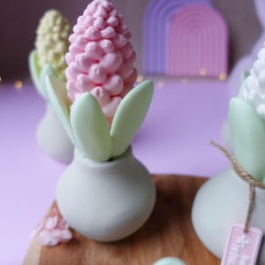 May include: A collection of decorative floral arrangements. Each arrangement features a pastel-colored flower, with a pink, yellow, and white flower, set in a white vase with green leaves. A wooden board and a purple background complete the scene.